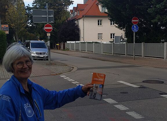 Eine Frau steht an einer Einbahnstraße und hält einen Flyer hoch, der auf die Gefahren des Linksradelns aufmerksam macht.