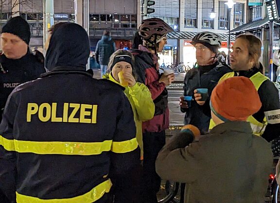 Frauen, Männer und Kinder stehen in einer Runde zusammen und unterhalten sich. Die meisten tragen Fahrradbekleidung. Im Vordergrund ist ein Polizist von hinten zu sehen.