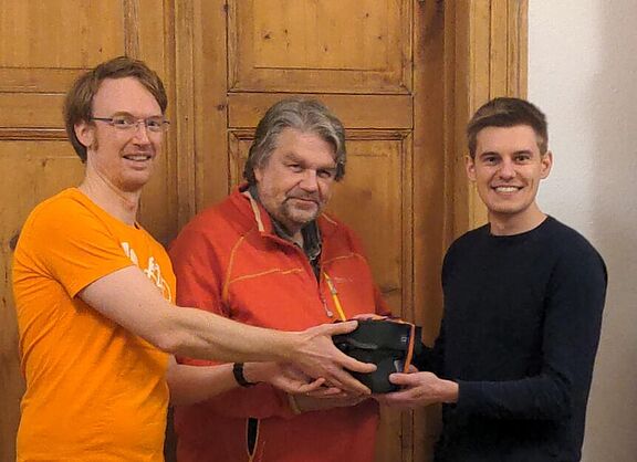 Drei Männer, von denen einer ein Geschenk überreicht bekommt.