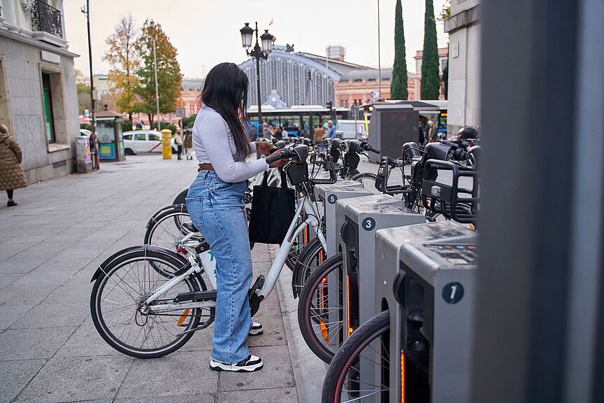 Frau entleiht Fahrrad an Bike-Sharing-Station Eine junge Frau mit langen dunklen Haaren, weißem Oberteil, Jeans und schwarzer Umhängetasche steht an einer Bike-Sharing-Station und entnimmt ein weißes Leihfahrrad. Die Station verfügt über mehrere nummerierte Stellplätze mit grauen Dockingelementen. Im Hintergrund sind ein belebter Stadtplatz, historische Gebäude, Passanten und ein Bahnhofsgebäude zu sehen.