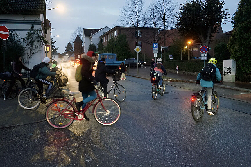 Junge Menschen fahren zur Schule Mehrere Jugendliche fahren auf dem Rad über eine Straße, während es morgens noch dämmert. Sie tragen Winterjacken und Rucksäcke, einige schützen ihren Kopf mit einem Helm. Die Lampen an den Rädern leuchten. Im Hintergrund warten Autos mit eingeschalteten Scheinwerfern an der Straße. Straßenlaternen erhellen die Szene.