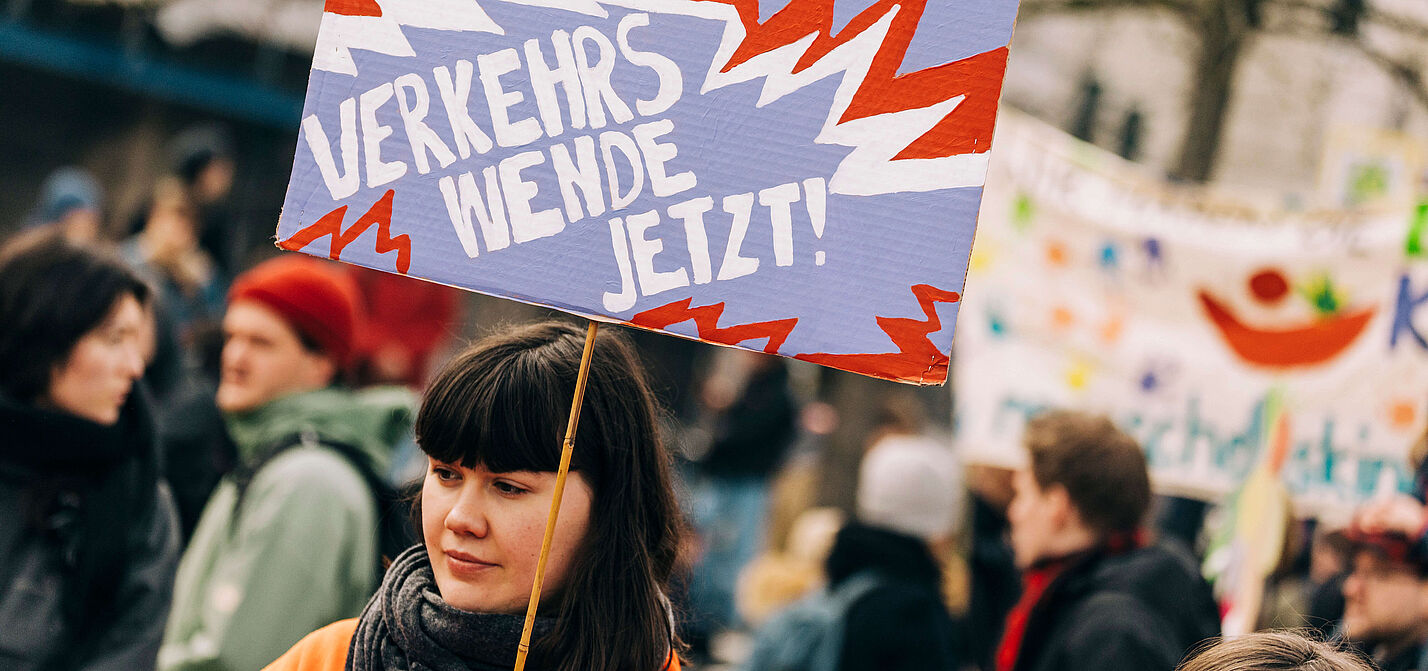 Junge Frau auf einer Demo hält ein Schild in die Luft, auf dem steht "Verkehrswende Jetzt!" 