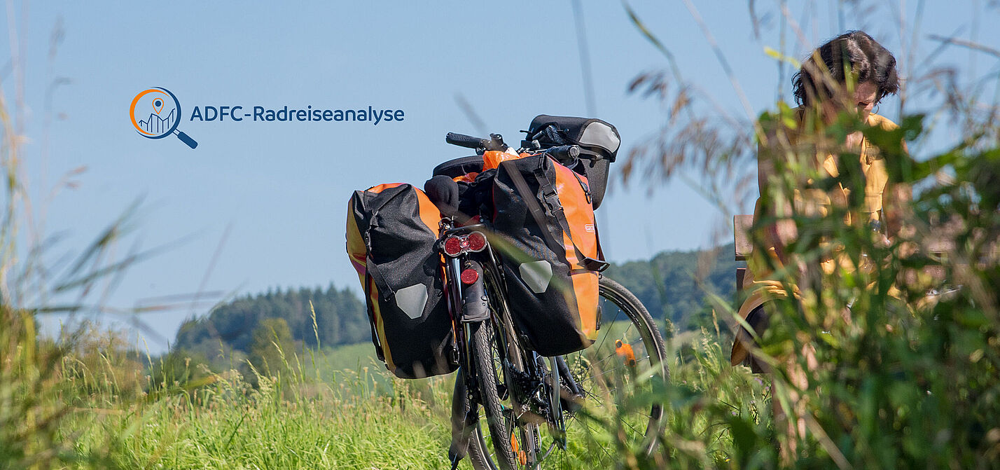 Reiserad mit Packtaschen steht in einer gemähten Wiese, Frau hinter hochgewachsenem Gras