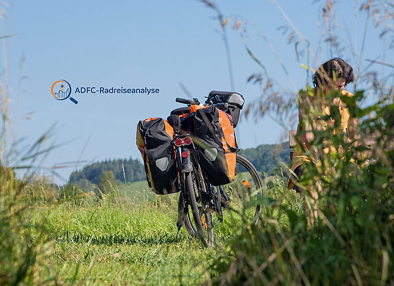 Reiserad mit Packtaschen steht in einer gemähten Wiese, Frau hinter hochgewachsenem Gras